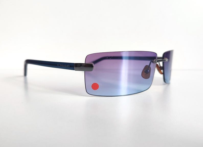 May include: Rimless sunglasses with gradient purple and blue lenses. The frames are a dark blue colour, with the brand name "Osse" printed on the temple. A small red dot is on the lower left lens.