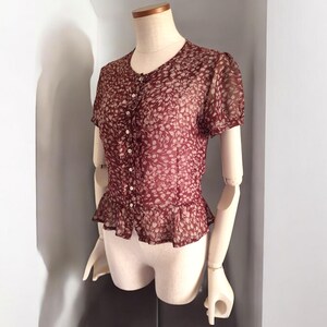 May include: A maroon sheer blouse with a white floral print and button-up front. The blouse has a ruffled hem and short sleeves.