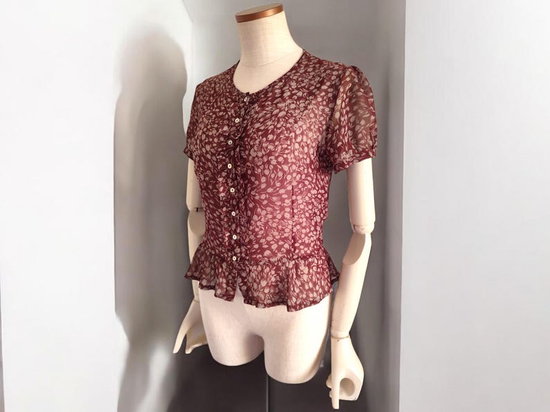 May include: A maroon sheer blouse with a white floral print and button-up front. The blouse has a ruffled hem and short sleeves.