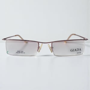 以下が含まれることがあります： Giada Luxのメガネ。フレームレスデザインで、フレームは薄茶色、レンズは長方形です。右レンズには「GIADA LUX」、左レンズには「MOD. 201 48□18-135 C4」と印字されています。