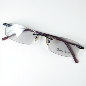 May include: Rimless eyeglasses with a burgundy frame and temples. The lenses are clear, and the brand name "SanMario" is printed on the right lens. The glasses have a sleek, modern design.