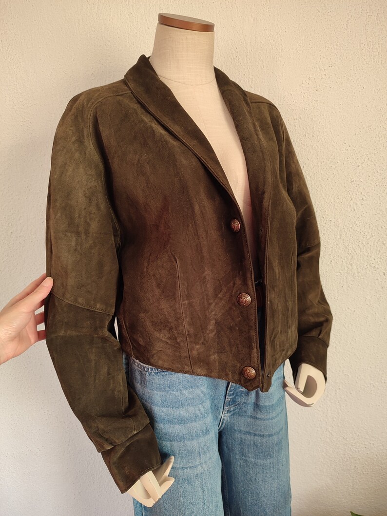 Puede incluir: Una chaqueta de ante marr&oacute;n con cuello y cierre de botones. La chaqueta tiene mangas largas y un dobladillo recortado.