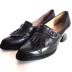 May include: A pair of black leather shoes with a low heel, a rounded toe, and a decorative fringe. The shoes feature a gold-colored buckle and a strap across the top. The shoes are set against a white background.