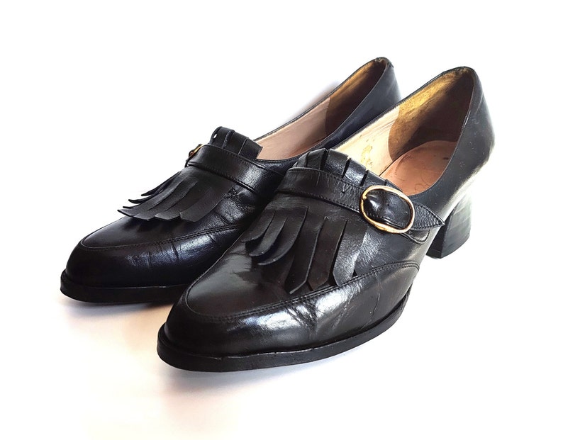 May include: A pair of black leather shoes with a low heel, a rounded toe, and a decorative fringe. The shoes feature a gold-colored buckle and a strap across the top. The shoes are set against a white background.