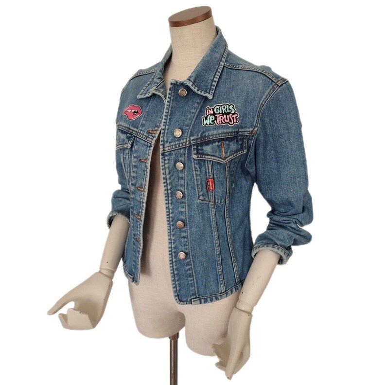 Pu&ograve; includere: Una giacca di jeans blu con una toppa rosa e verde che dice "In Girls We Trust" e una toppa di labbra rosse.