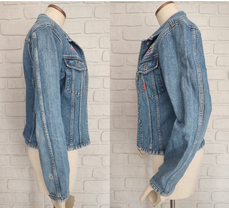 Pu&ograve; includere: Una giacca di jeans blu chiaro con una toppa rossa e bianca sul lato sinistro e una toppa rossa e bianca sul lato destro. La giacca ha una chiusura con bottoni e maniche lunghe.