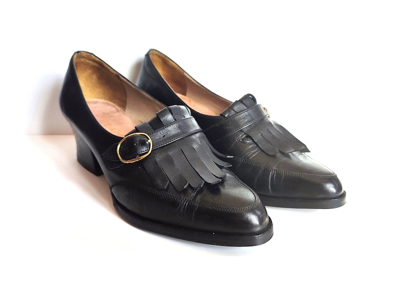 May include: A pair of black leather shoes with a block heel. The shoes feature a strap with a gold-colored buckle and decorative fringe. The shoes are displayed on a white background.