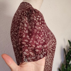 May include: A maroon sheer blouse with a white floral print. The blouse has a round neckline and short sleeves with a gathered detail at the cuff.
