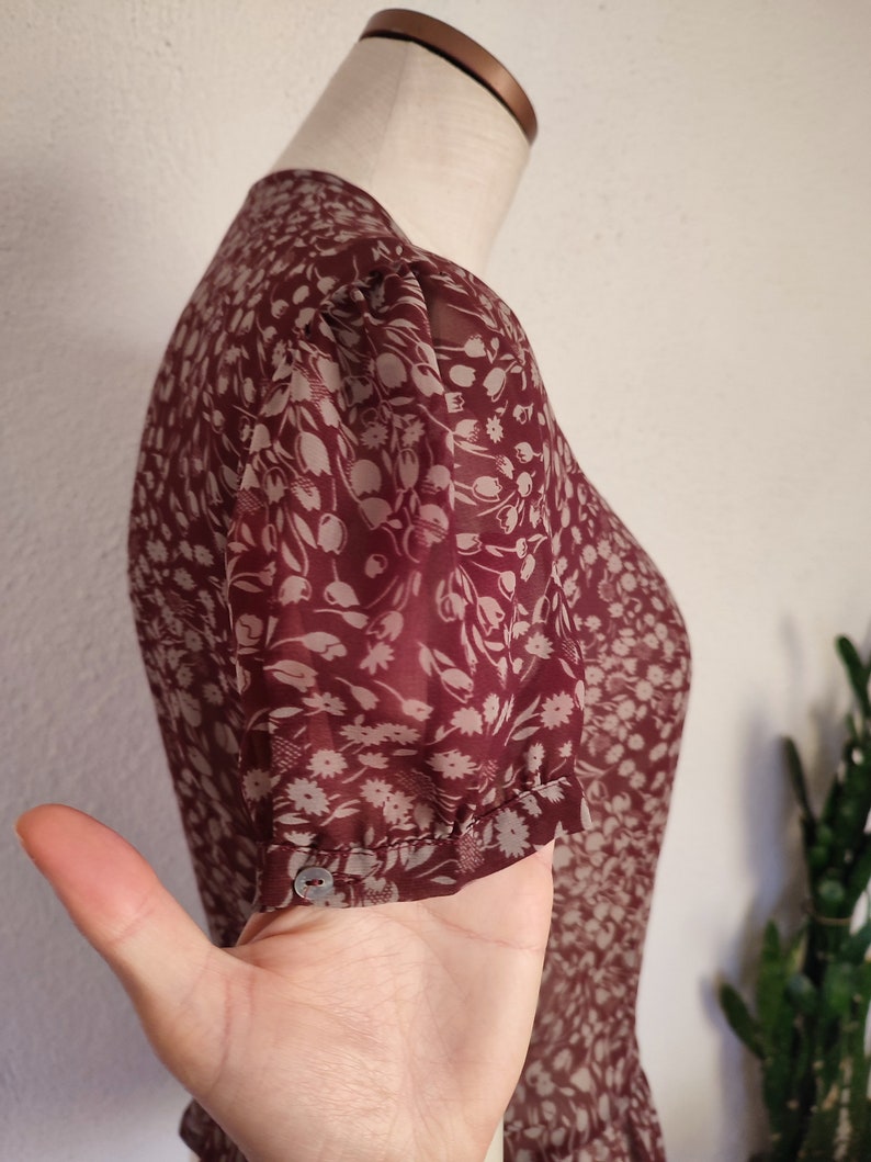 May include: A maroon sheer blouse with a white floral print. The blouse has a round neckline and short sleeves with a gathered detail at the cuff.