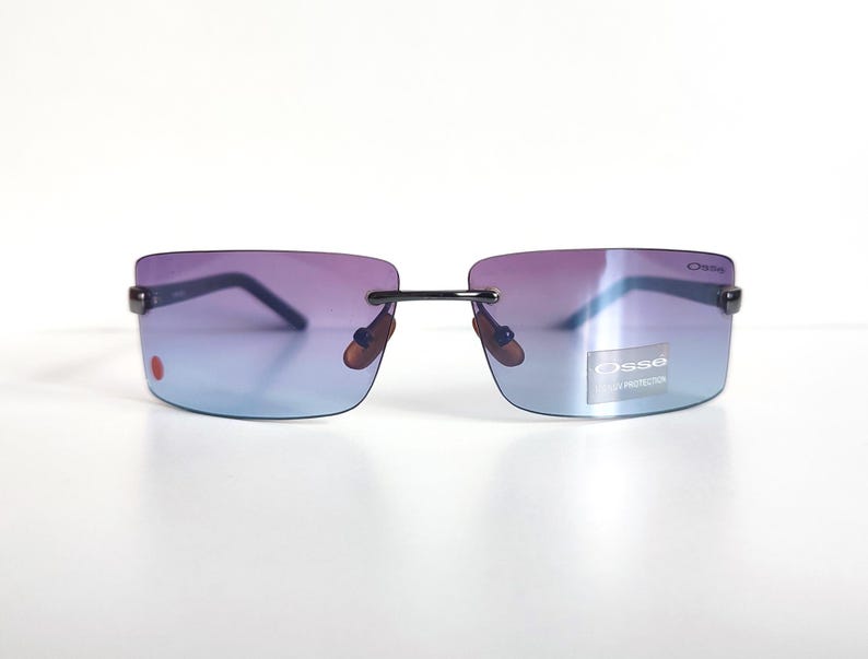May include: Rimless sunglasses with a gradient lens, shifting from purple to blue. The sunglasses have a black frame and the brand name "Osse" is visible on the lens. A small red dot is on the left lens.
