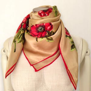 Könnte beinhalten: Ein beigefarbener Seidenschal mit einem auffälligen Blumenmuster. Der Schal zeigt große rote Blumen mit grünen Blättern und einen roten Rand. Der Schal ist über eine Schaufensterpuppe drapiert, um seine Form und sein Design hervorzuheben. Der Schal ist quadratisch.