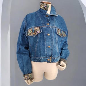 May include: A blue denim jacket with gold and green embroidered trim on the collar, pockets, and cuffs.