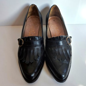 May include: A pair of black leather loafers with a pointed toe and a small gold-colored buckle. The shoes have a fringe detail across the top and a low heel. The interior of the shoe is a light tan color.