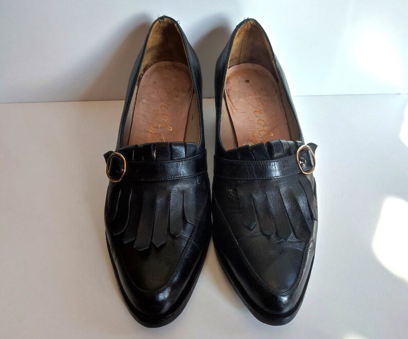 May include: A pair of black leather loafers with a pointed toe and a small gold-colored buckle. The shoes have a fringe detail across the top and a low heel. The interior of the shoe is a light tan color.