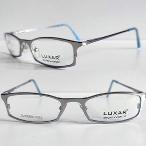 Può includere: Occhiali da vista in metallo argentato con montatura rettangolare. Le aste sono blu e le lenti sono trasparenti. Il nome del marchio "LUXAR eyewear" è stampato sulla montatura.