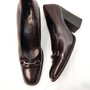 May include: A pair of brown leather heeled pumps with a silver buckle detail. The shoes have a square toe and a chunky heel.