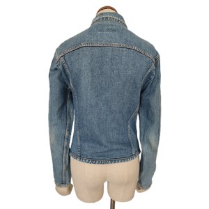 Pu&ograve; includere: Una giacca di jeans blu con chiusura a bottoni e maniche lunghe. La giacca &egrave; su un manichino bianco.