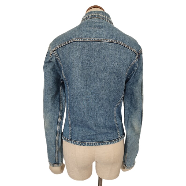 Pu&ograve; includere: Una giacca di jeans blu con chiusura a bottoni e maniche lunghe. La giacca &egrave; su un manichino bianco.