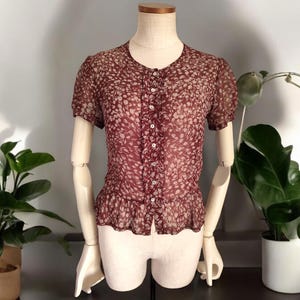 May include: A maroon sheer blouse with a white floral pattern and a button-up front. The blouse has a ruffled hem and short sleeves.