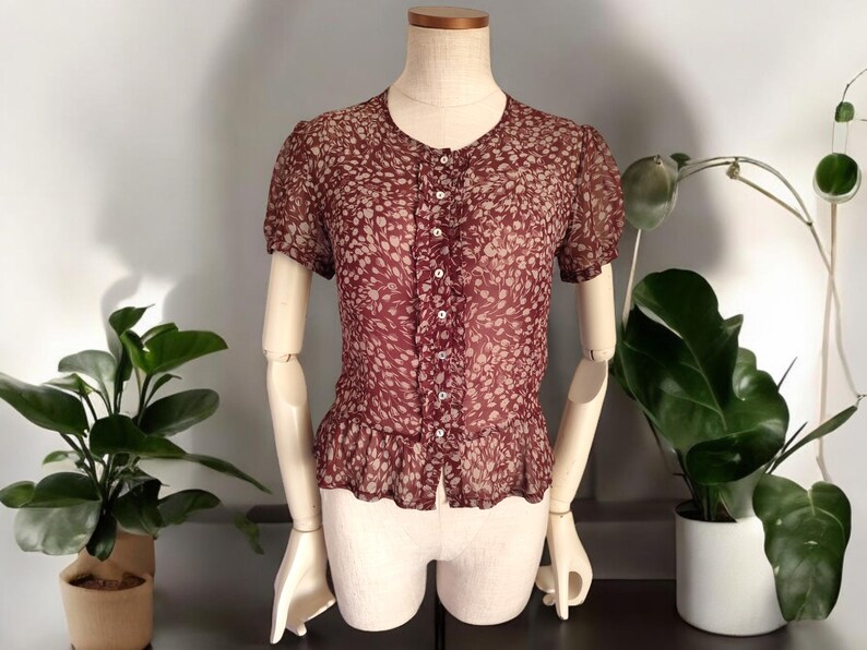 May include: A maroon sheer blouse with a white floral pattern and a button-up front. The blouse has a ruffled hem and short sleeves.