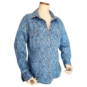 May include: A long-sleeved denim shirt with a light blue paisley pattern. The shirt has a classic collar, two chest pockets, and silver snap closures. The cuffs also have snap closures. The shirt is displayed on a mannequin.