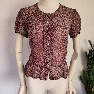 May include: A maroon sheer blouse with a white floral print and a button-up front. The blouse has short puffed sleeves and a ruffled hem.