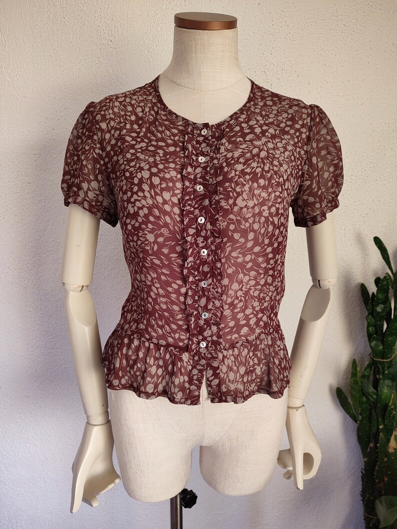 May include: A maroon sheer blouse with a white floral print and a button-up front. The blouse has short puffed sleeves and a ruffled hem.