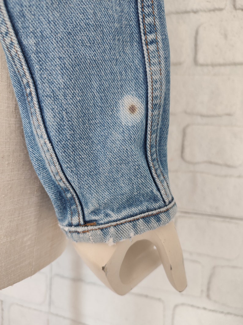 Pu&ograve; includere: Primo piano della manica in denim blu chiaro di una giacca. La manica presenta una piccola macchia marrone.