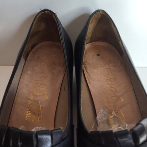 May include: A pair of black leather pumps with a low heel. The shoes have a decorative pleated detail across the vamp and a gold-colored buckle. The insoles are tan with gold lettering.