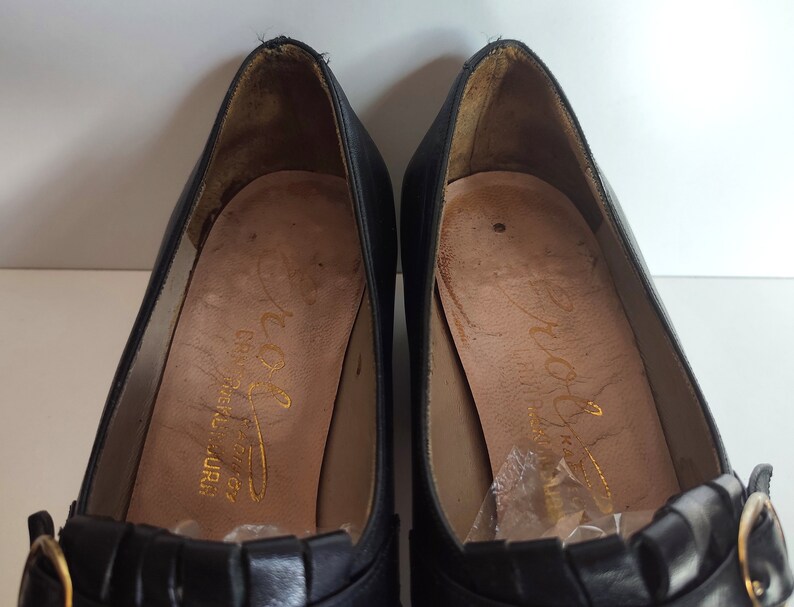 May include: A pair of black leather pumps with a low heel. The shoes have a decorative pleated detail across the vamp and a gold-colored buckle. The insoles are tan with gold lettering.