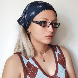 Op de afbeelding: Een marineblauwe paisley bandana gedragen als hoofdbedekking. De persoon draagt een zwarte rechthoekige bril en een bruin en blauw argyle patroon tanktop. Een gouden ketting met een kleine hanger is ook zichtbaar.