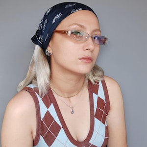 May include: A person wearing a navy blue bandana with a paisley design, silver-rimmed glasses, and a brown and blue argyle patterned tank top. The person is also wearing a silver necklace and hoop earrings.