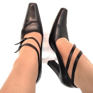 May include: A pair of black leather heeled shoes with double straps and buckles. The shoes are worn on a person's feet.