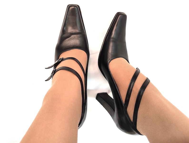 May include: A pair of black leather heeled shoes with double straps and buckles. The shoes are worn on a person's feet.