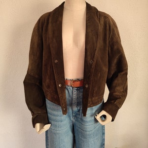 Puede incluir: Una chaqueta de ante marr&oacute;n con cuello y cierre de botones. La chaqueta es corta y tiene un corte holgado. Se muestra en un maniqu&iacute; que lleva pantalones vaqueros azules y un cintur&oacute;n marr&oacute;n.