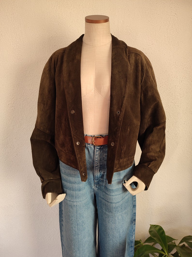 Puede incluir: Una chaqueta de ante marr&oacute;n con cuello y cierre de botones. La chaqueta es corta y tiene un corte holgado. Se muestra en un maniqu&iacute; que lleva pantalones vaqueros azules y un cintur&oacute;n marr&oacute;n.
