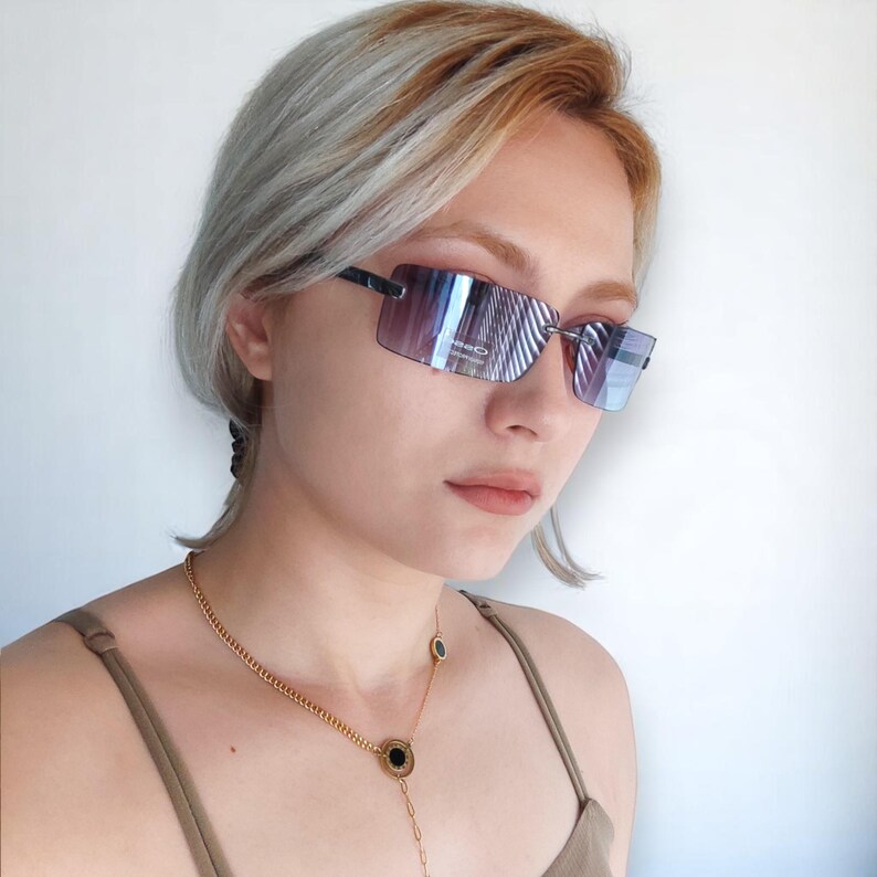 May include: Rimless sunglasses with a gradient lens and a rectangular shape, set in a dark frame. A gold chain necklace with black circular pendants is also visible. The person is wearing a brown top.