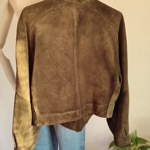 Puede incluir: Una chaqueta de ante verde oscuro con cuello y mangas largas. La chaqueta tiene cierre de botones y un dobladillo ligeramente recortado.