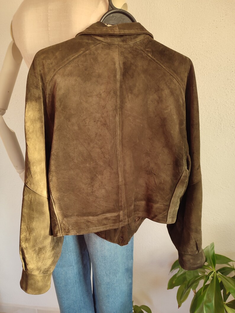 Puede incluir: Una chaqueta de ante verde oscuro con cuello y mangas largas. La chaqueta tiene cierre de botones y un dobladillo ligeramente recortado.