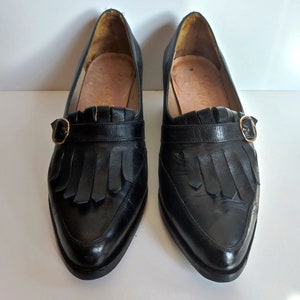 May include: A pair of black leather loafers with a pointed toe, fringe detail, and a gold-colored buckle. The shoes have a low heel and a leather sole. The interior of the shoe is a light brown color.