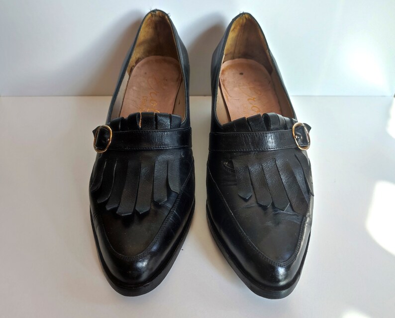 May include: A pair of black leather loafers with a pointed toe, fringe detail, and a gold-colored buckle. The shoes have a low heel and a leather sole. The interior of the shoe is a light brown color.