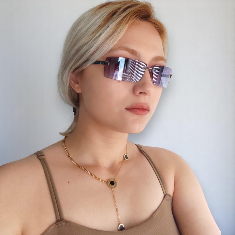 May include: Rimless sunglasses with a gradient purple tint and black frames. A gold necklace with black circular pendants is also visible. The model is wearing a brown vest top.