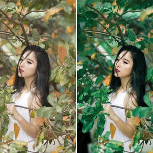 May include: A woman with long black hair stands in front of a leafy green tree. She is wearing a white sleeveless top and has a branch in her hand.