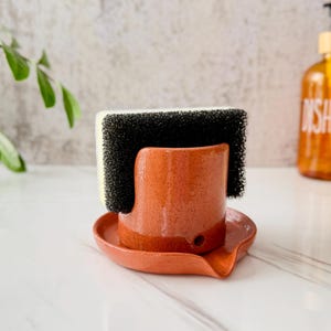May include: A terracotta-coloured ceramic sponge holder with a glossy finish and a small drainage hole at the base, featuring a curved design that holds a rectangular yellow and black scouring sponge upright within its integrated circular tray.