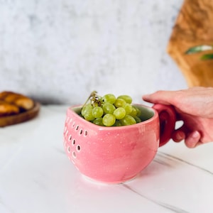 May include: A pink ceramic berry bowl with a rounded shape and a handle on the side. The bowl features a series of small, circular drainage holes on its exterior surface and is filled with a cluster of fresh, green grapes.