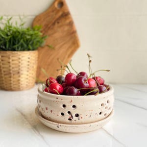 May include: A ceramic berry bowl filled with fresh cherries. The bowl is off-white with small holes and a matching saucer. The cherries are a mix of red and dark burgundy, with green stems. A small potted plant and wooden cutting board are in the background.
