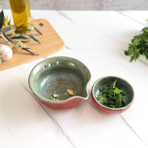 May include: A ceramic kitchen set consisting of a larger green and terracotta bowl with a textured interior and a pouring spout, alongside a smaller matching bowl filled with fresh green parsley leaves, both featuring a smooth glazed finish.
