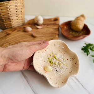 May include: A speckled, off-white ceramic garlic grater with a scalloped edge, holding grated garlic and a clove. A wooden cutting board, garlic bulb, and mortar and pestle are in the background. Kitchenware.