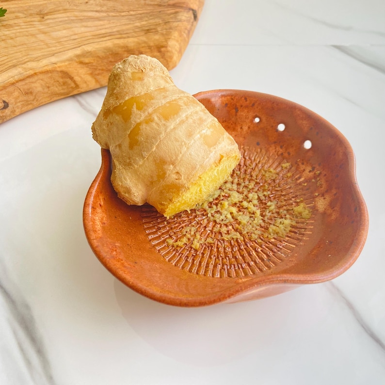 May include: A fresh ginger root rests on a terracotta ginger grater. The grater has a textured surface for efficient grating. The ginger is a light brown color, and the grater is a warm reddish-brown. A wooden cutting board is in the background.