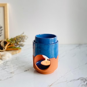 Peut inclure: Un brûleur à huile en céramique bleu et terracotta avec un glaçage turquoise. Le brûleur a une ouverture ronde sur le côté pour accueillir une bougie chauffe-plat. La bougie est allumée et la flamme est visible.